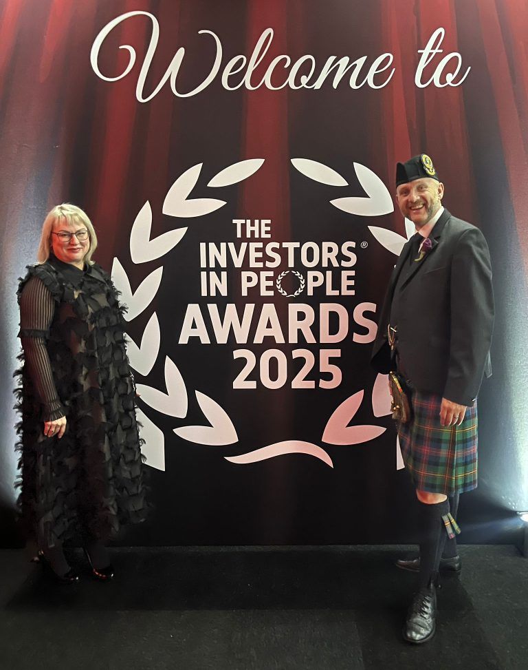 A man and woman smartly dressed at an awards ceremony stand in front of a photo background for the IIP Awards 2025 Nomination