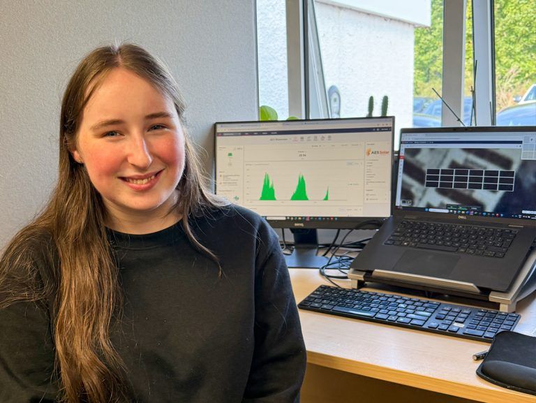 A young woman sits at a desk facing the camera on work experience at aes renewables with two computer screens in the background.