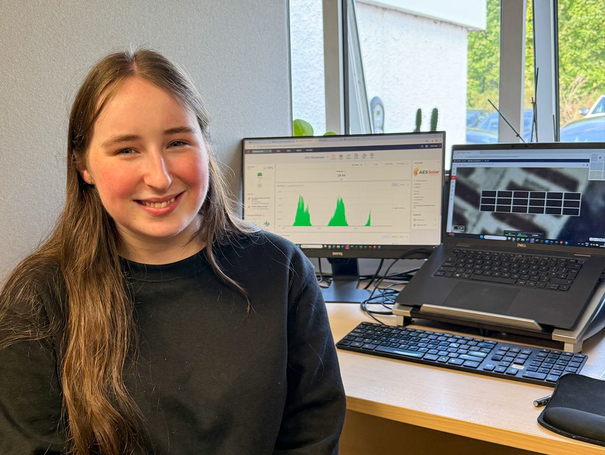 A young woman sits at a desk facing the camera on work experience at aes renewables with two computer screens in the background.